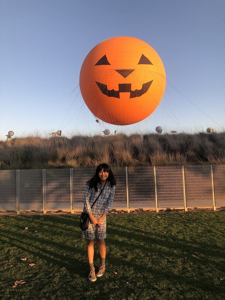 Eva is smiling and standing in a grassy field in front of a big hot air balloon that looks like a jack-o-lantern.
